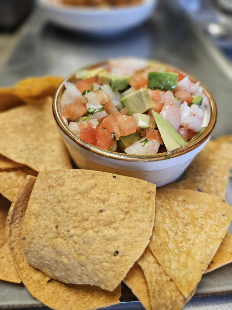 Ceviche: "A colorful dish of fresh ceviche featuring marinated shrimp, diced tomatoes, onions, cilantro, and avocado, served with crisp tortilla chips on the side."